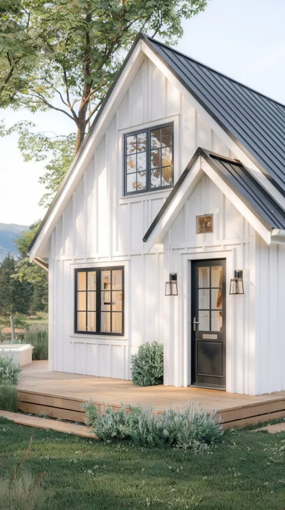 A modern white Scandinavian cottage with vertical siding, crisp black-framed windows, and a dark metal roof, featuring a small covered entrance and a wooden deck, set against a backdrop of trees and a mountain.