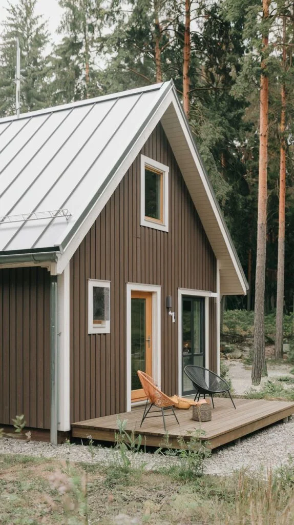 A contemporary dark brown Scandinavian cottage with vertical siding, white-framed windows, and a light-colored metal roof, featuring a prominent light wooden front door and a deck with outdoor seating.