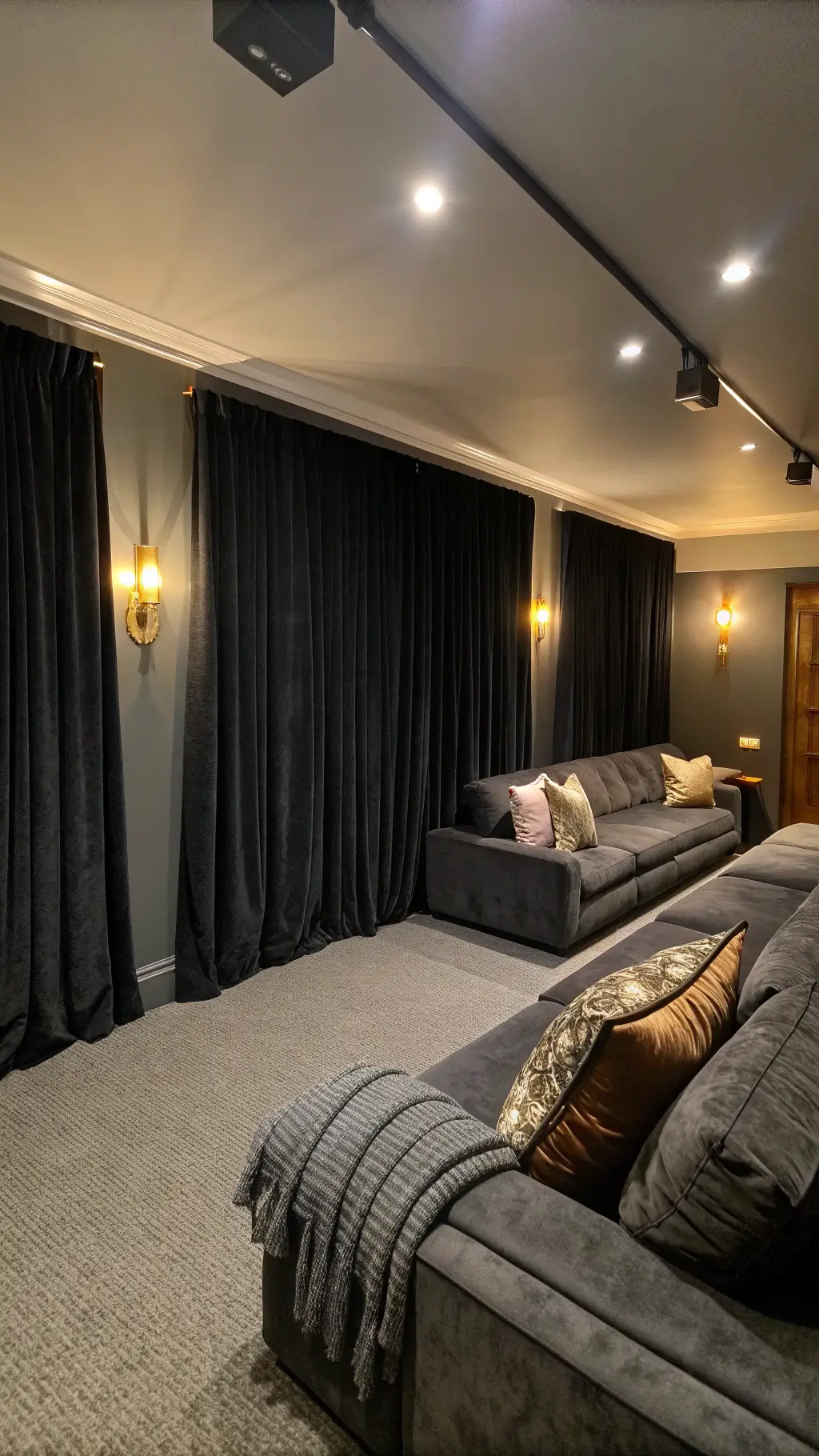 Cozy media room with automated double-layer black curtains, charcoal sectional, textured pillows, and brass wall sconces creating evening ambiance.