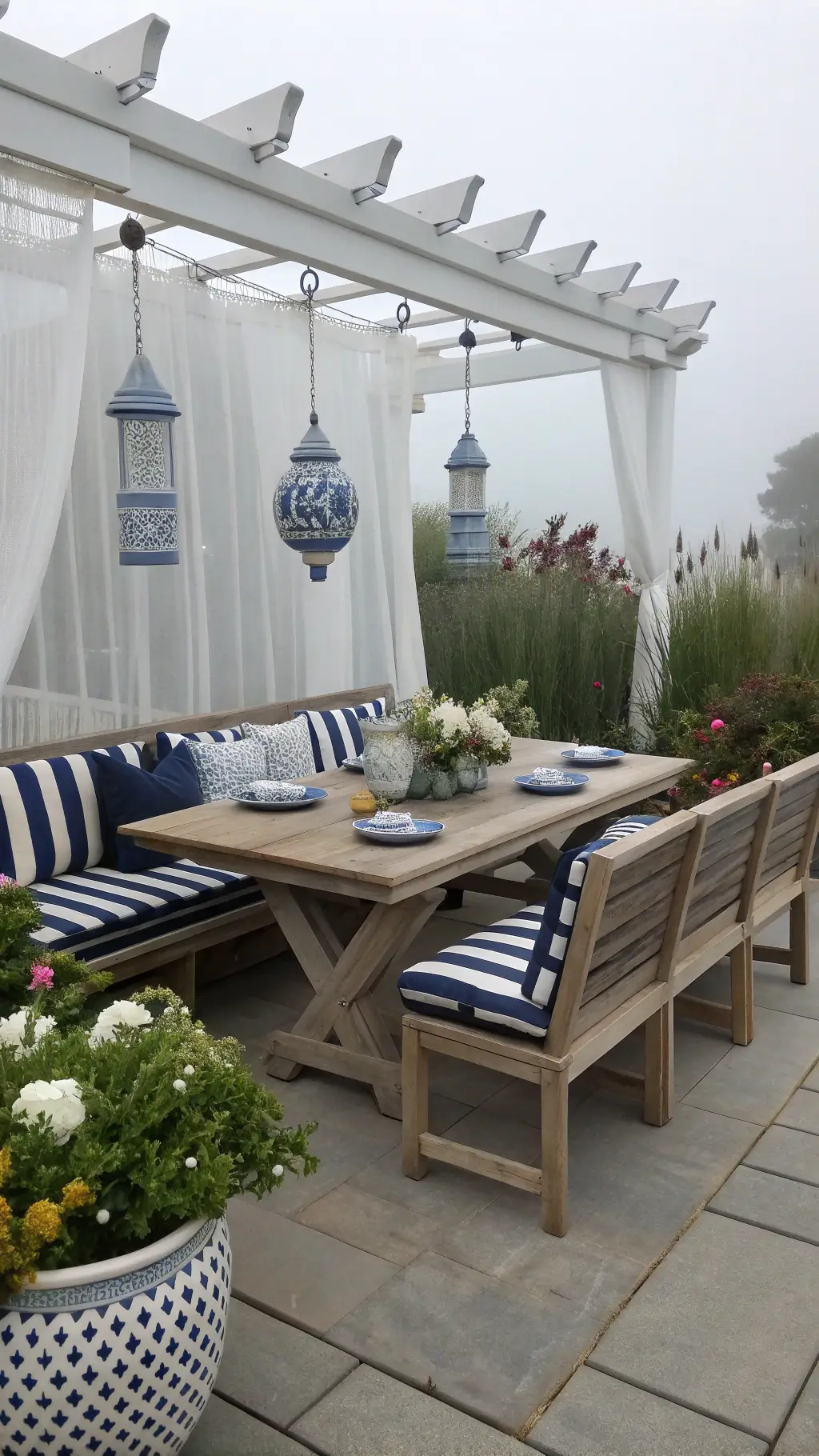 Teak dining set on a coastal-inspired patio with white lattice backdrop, jasmine drapes, blue ceramics, and marine-style pendant lights creating a serene atmosphere