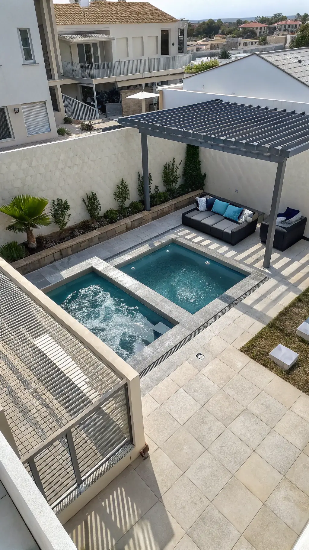 Overhead view of a modern gray prefabricated cocktail pool with geometric design, corner spa, sand-colored porcelain tiles, charcoal sectional seating with blue accents under a steel pergola, enclosed by white privacy screens with climbing jasmine