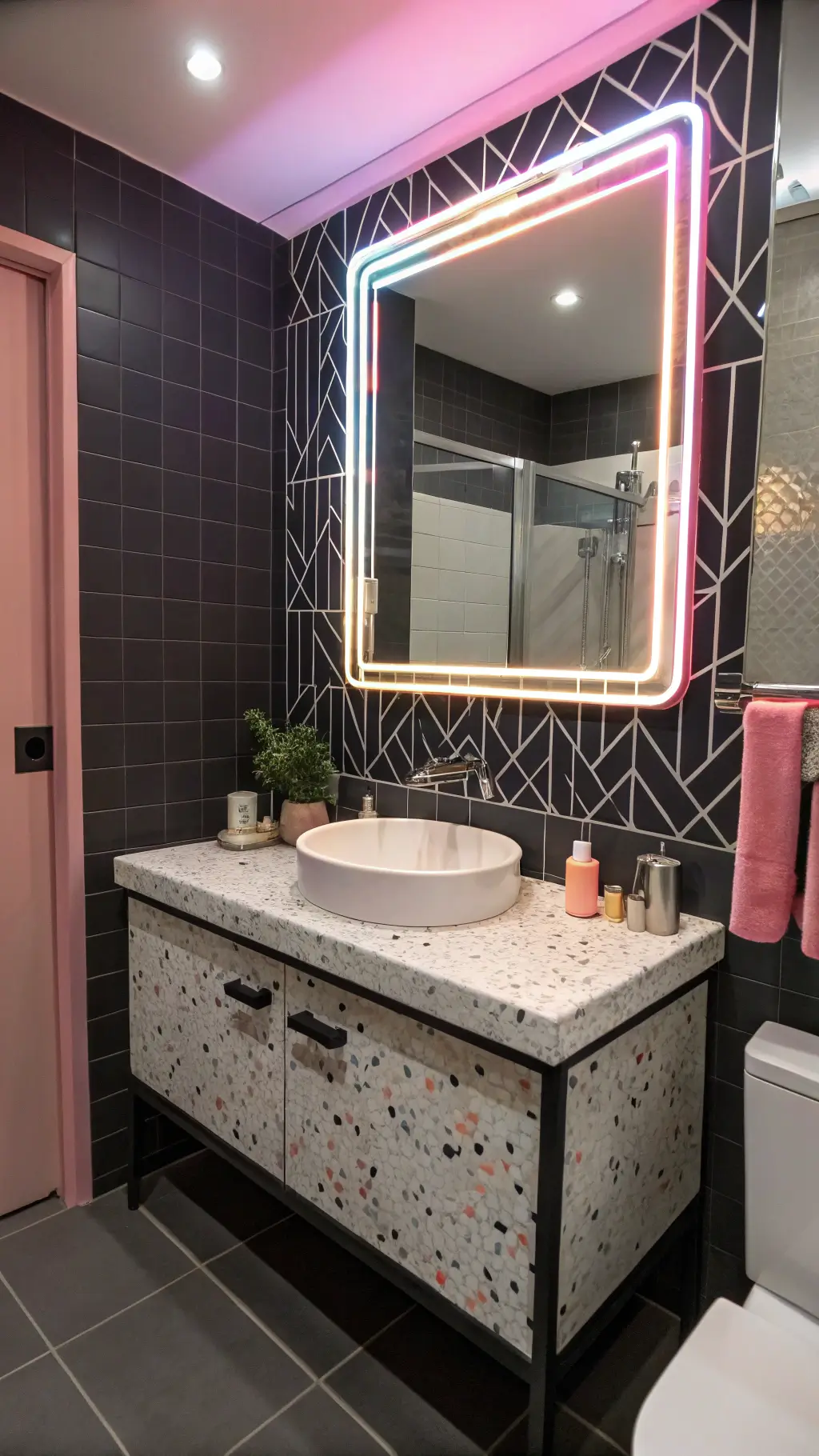 Bathroom vanity with floating chrome base and pastel terrazzo top, neon pink Memphis-style mirror border, and matte black geometric tiles, accentuated by dramatic side lighting.