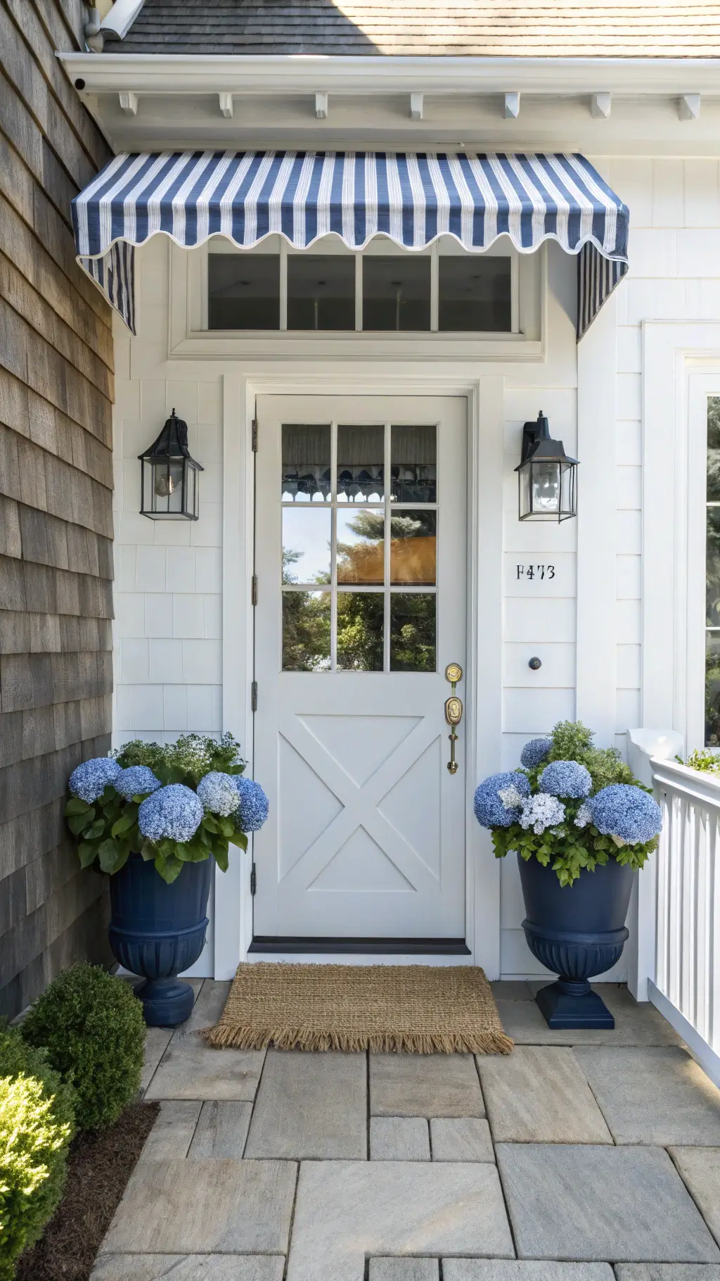 Bright sunlit coastal Hamptons entryway with white Dutch door featuring X-pattern, navy and striped awning, brass marine-style sconces, blue hydrangeas in ceramic planters, natural sisal doormat on bluestone porch
