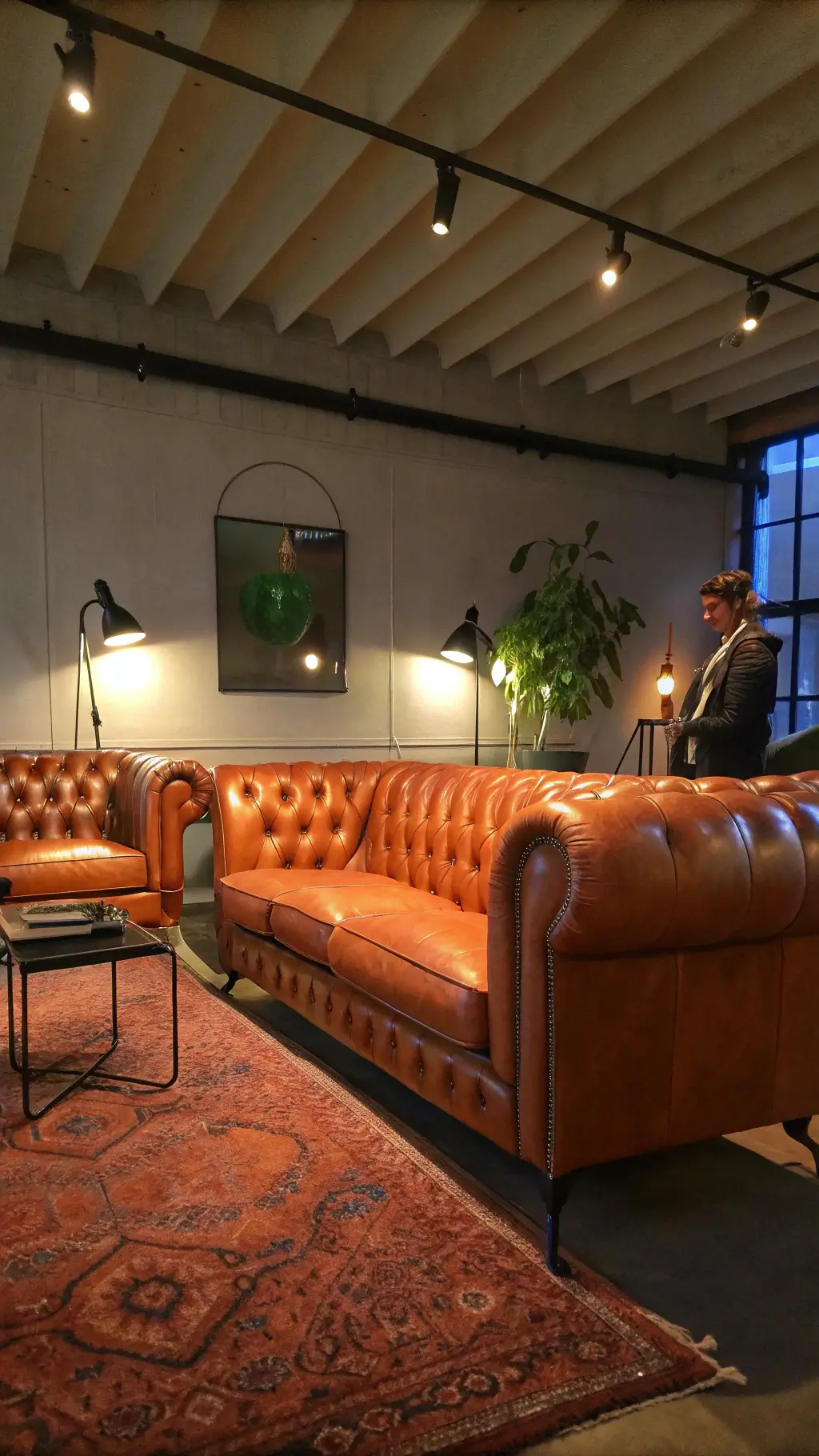 vintage-inspired living room with classic chesterfield sofa, modern steel-framed chairs, layered rugs, black metal sconces, textured walls under moody evening lighting