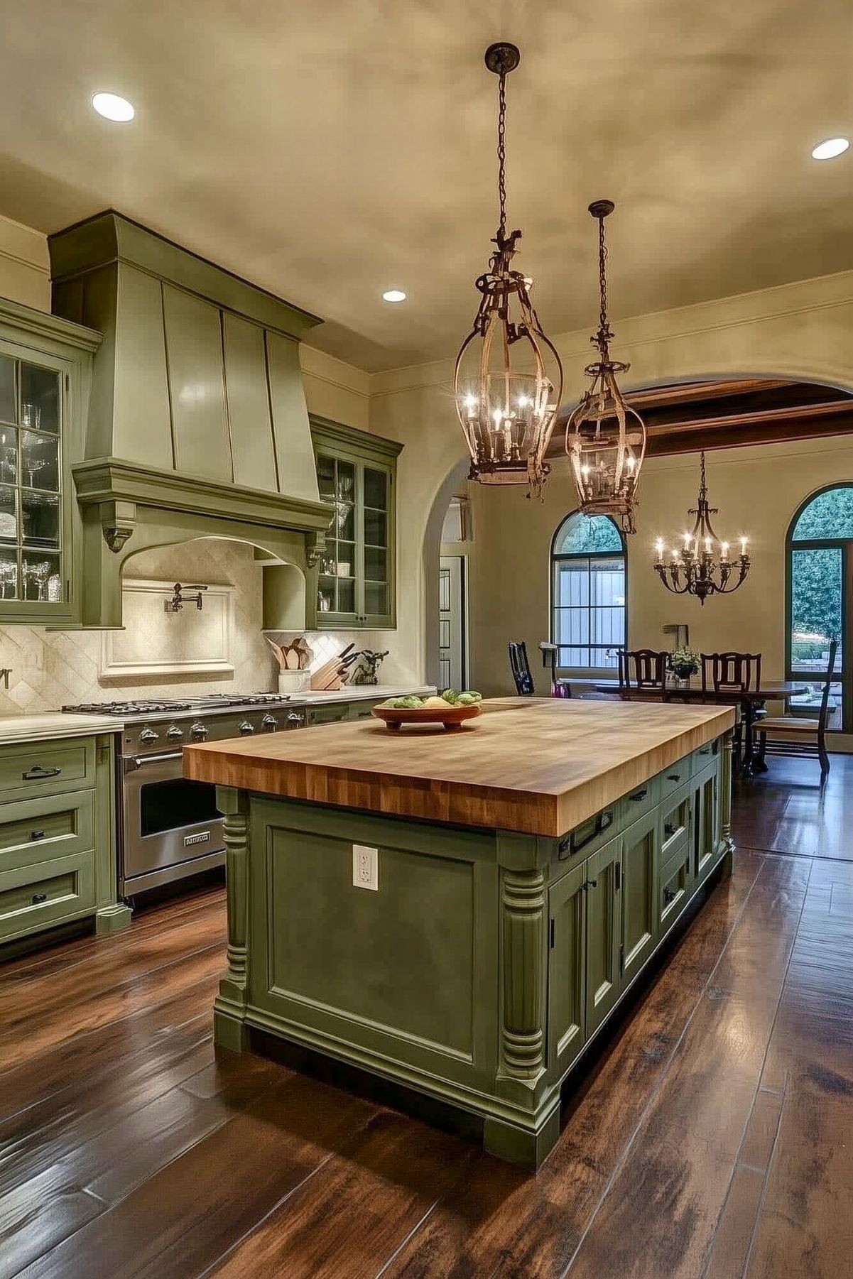 Classic kitchen with olive green cabinets, large wooden island, ornate chandeliers, and warm wood flooring, creating a welcoming traditional atmosphere.